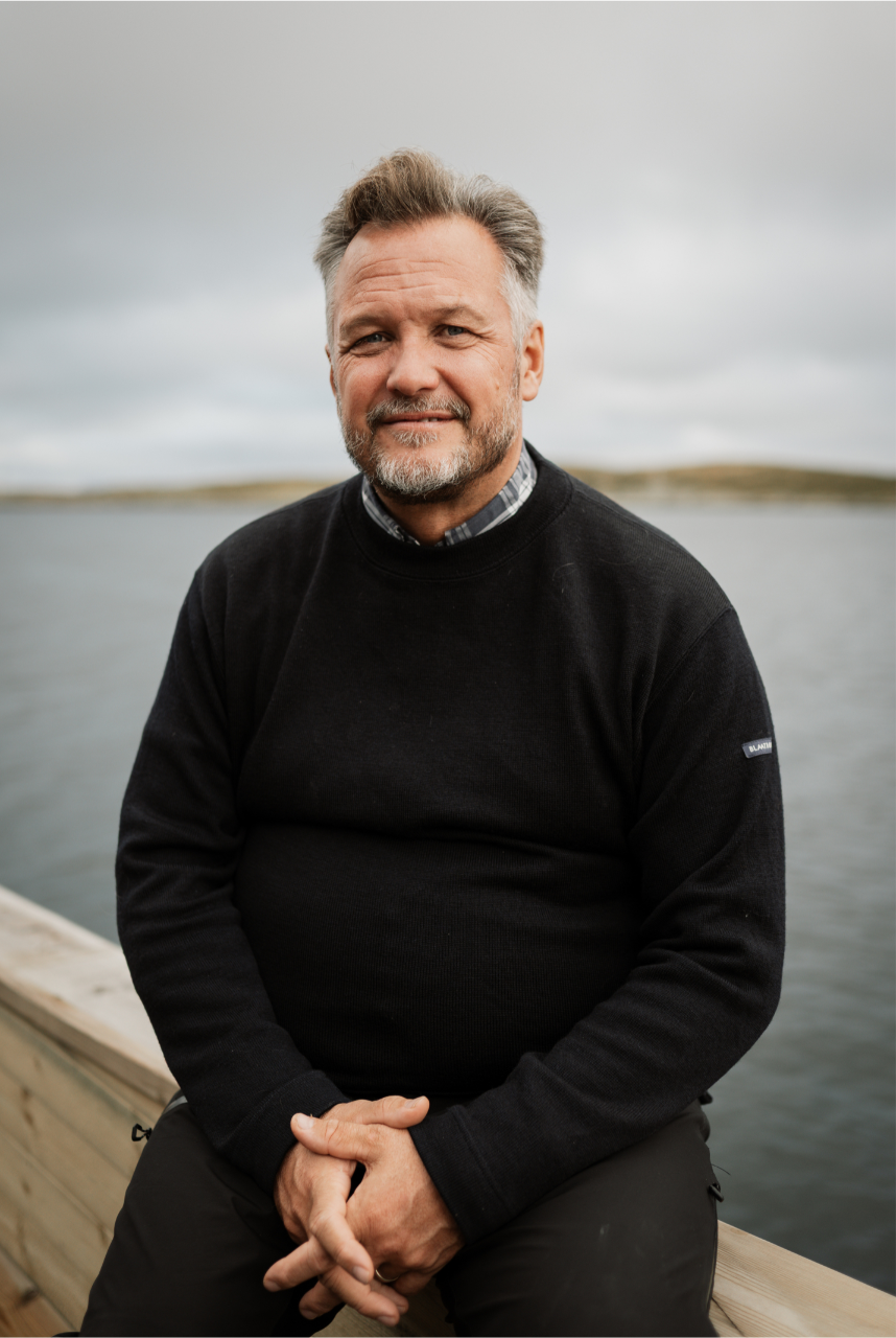 Mikael Forselius VISIT Group. A person in a dark sweater sits on a wooden dock by calm water under an overcast sky, face blurred.