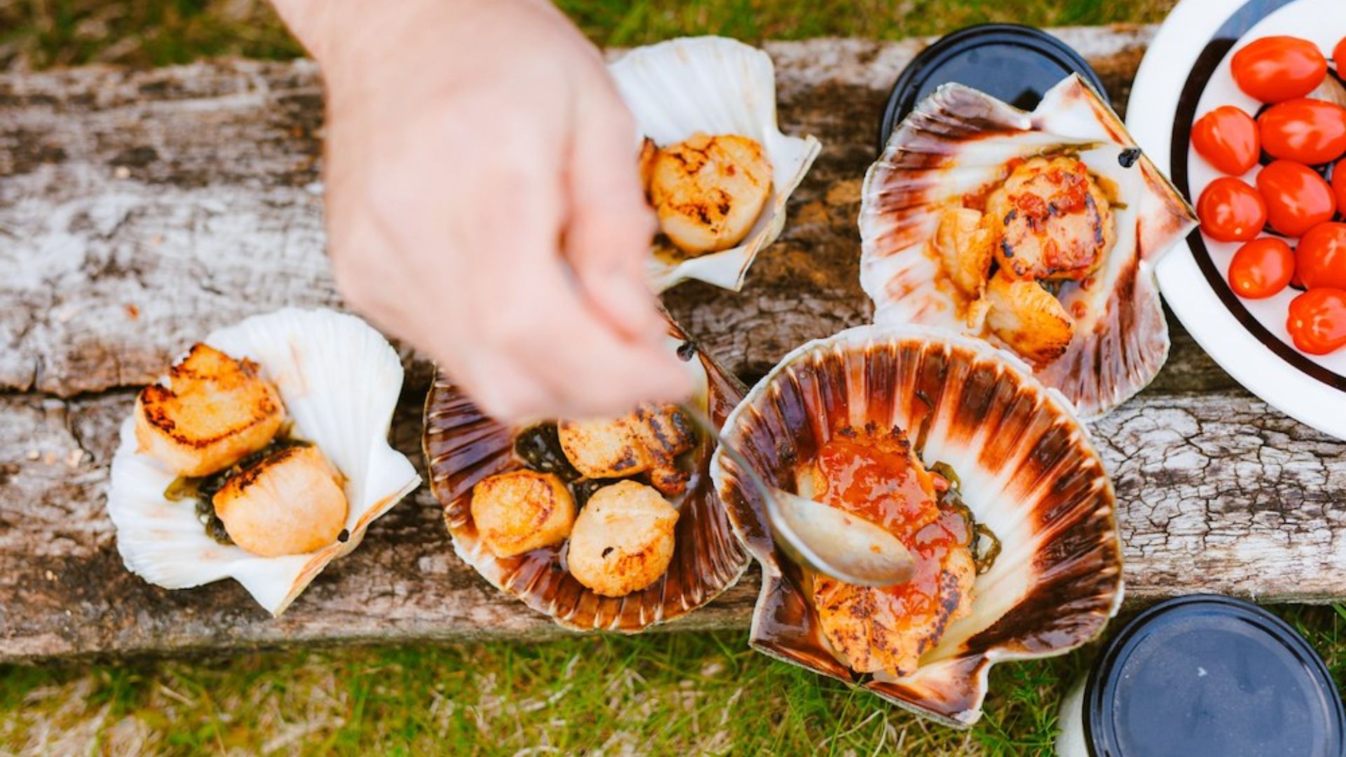 Hand adds sauce to grilled scallops served in scallop shells on a rustic wooden picnic table with tomatoes.
