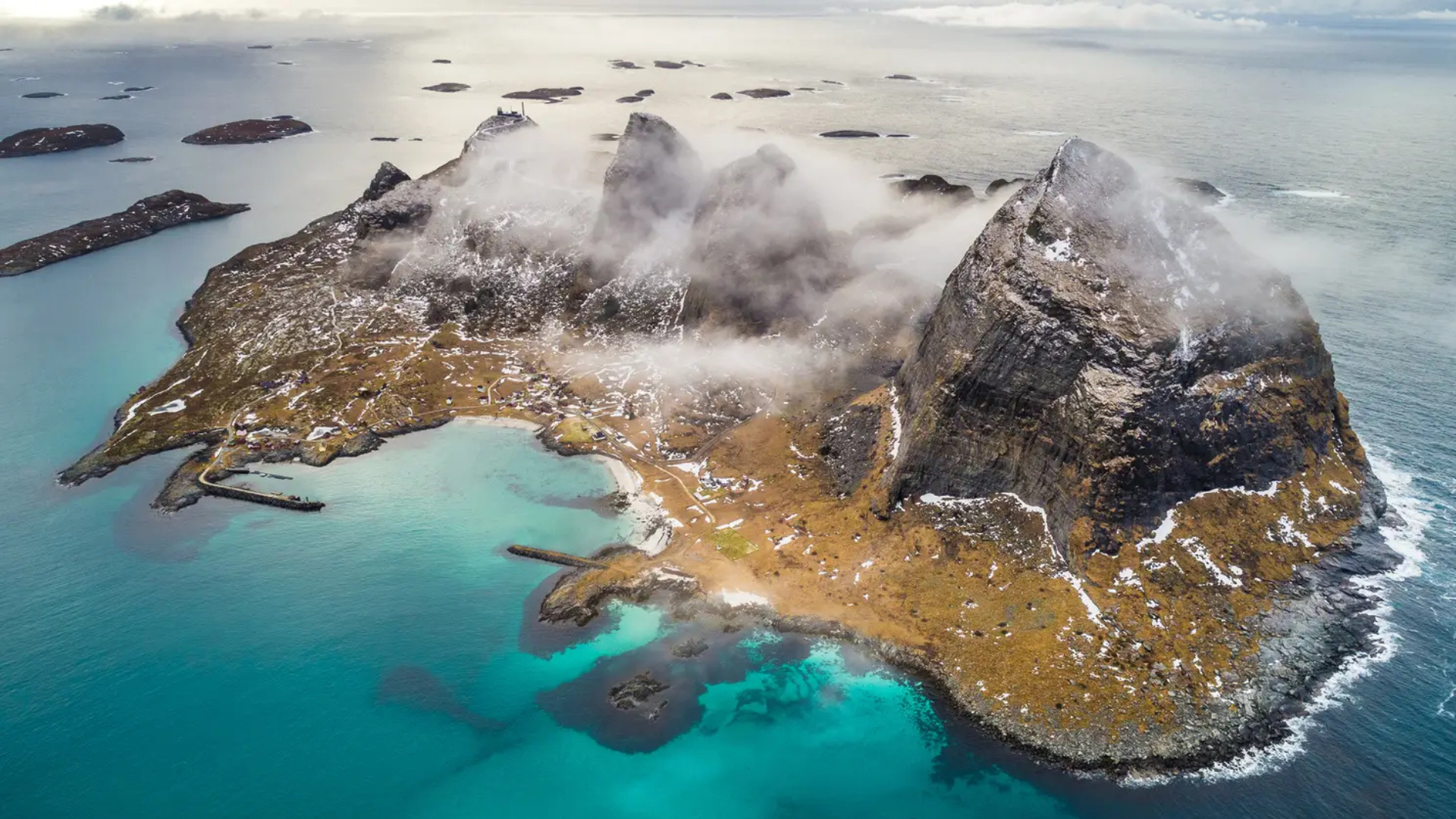 Aerial view of a rugged island with steep mountains in low cloud, patches of snow, and bright turquoise coastal water.