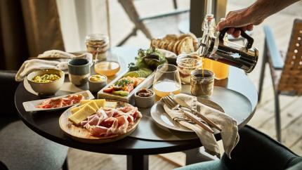 A hand pours coffee into a mug beside a table set with bread, cheese, cured meat, fruit, and juice.