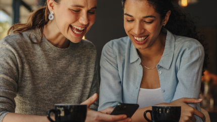 Two people sit at a cafe table with coffee cups, one holding a smartphone, with warm indoor lighting behind them.