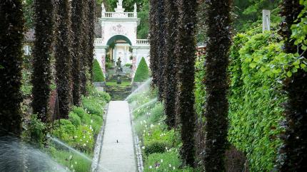 Unike Hoteller and VISIT Partnership_Tree-lined garden walkway with sprinklers watering flowerbeds, leading toward an ornate white archway gate.