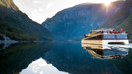 Modern boat cruising through a calm fjord with mountains and sunlight in the background.