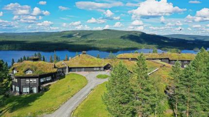 Aerial view of grass-roofed buildings on a hillside above a lake, with forested mountains and clouds beyond.