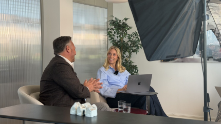 Two people talk in a modern office lounge, with a laptop on a side table and studio lighting equipment in view.