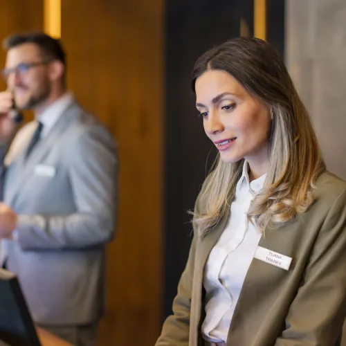 woman working in hotel reception, man in the background answering the phone