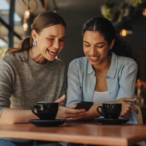 Two people sit at a cafe table with coffee cups, one holding a smartphone, with warm indoor lighting behind them.