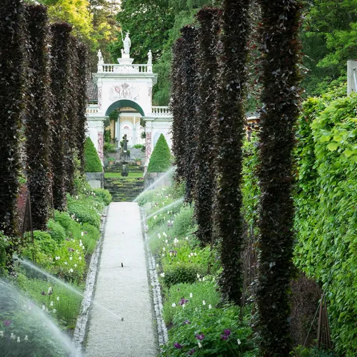Unike Hoteller and VISIT Partnership_Tree-lined garden walkway with sprinklers watering flowerbeds, leading toward an ornate white archway gate.