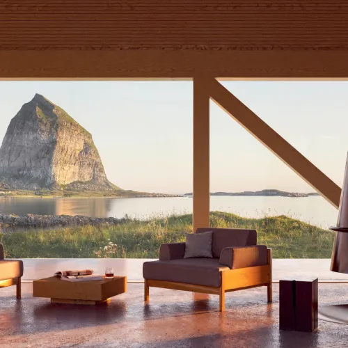 Modern lounge with two armchairs and a suspended fireplace, facing a calm bay and a steep mountain at sunset.