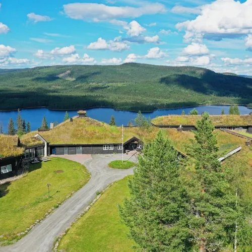 Aerial view of grass-roofed buildings on a hillside above a lake, with forested mountains and clouds beyond.