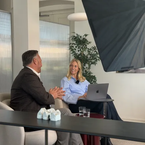 Two people talk in a modern office lounge, with a laptop on a side table and studio lighting equipment in view.